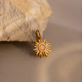 A close-up image of a gold Opal Sun Charm resting against a textured beige stone. The charm features a radiant, sculpted sunburst design with an iridescent opal stone at its center, glowing with hues of orange, green, and blue. The charm has a small polished gold bail for attachment.