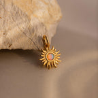 A close-up image of a gold Opal Sun Charm resting against a textured beige stone. The charm features a radiant, sculpted sunburst design with an iridescent opal stone at its center, glowing with hues of orange, green, and blue. The charm has a small polished gold bail for attachment.
