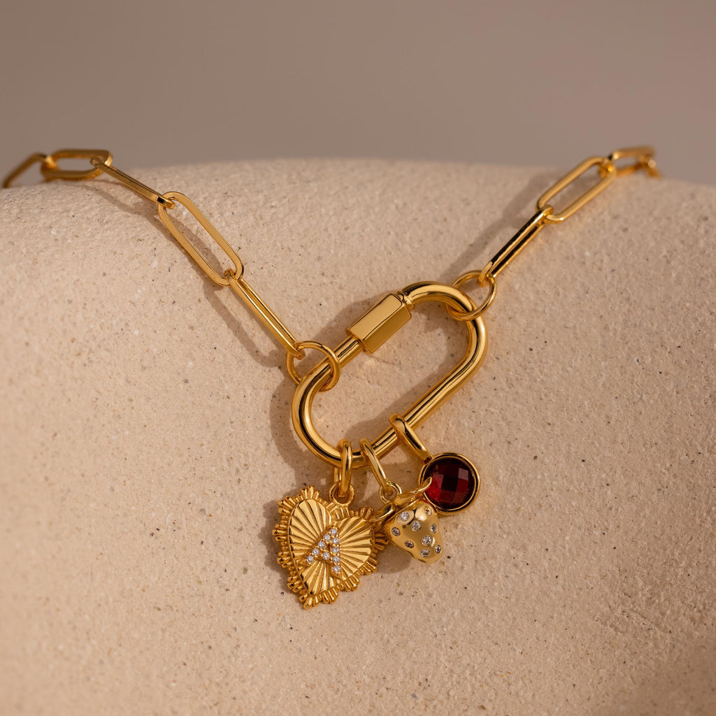 A gold Ribbed Heart Initial Pendant with a pavé letter is styled on a carabiner clasp link necklace alongside a strawberry charm and a red garnet gemstone charm