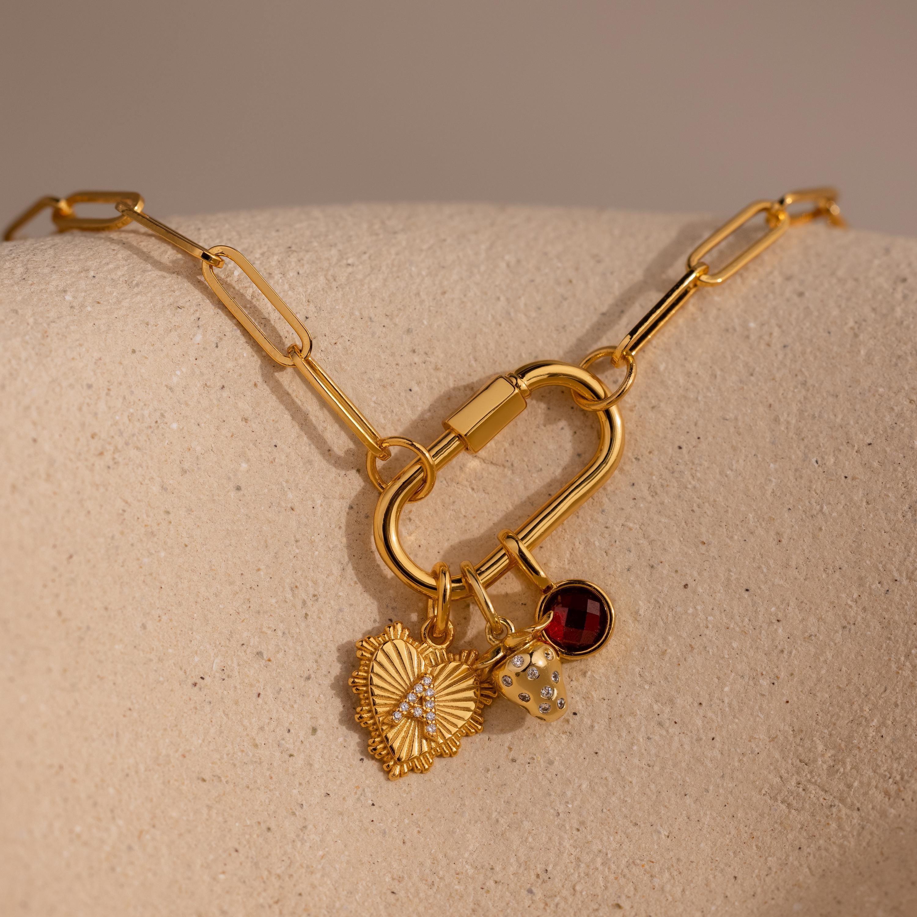 A gold Ribbed Heart Initial Pendant with a pavé letter is styled on a carabiner clasp link necklace alongside a strawberry charm and a red garnet gemstone charm