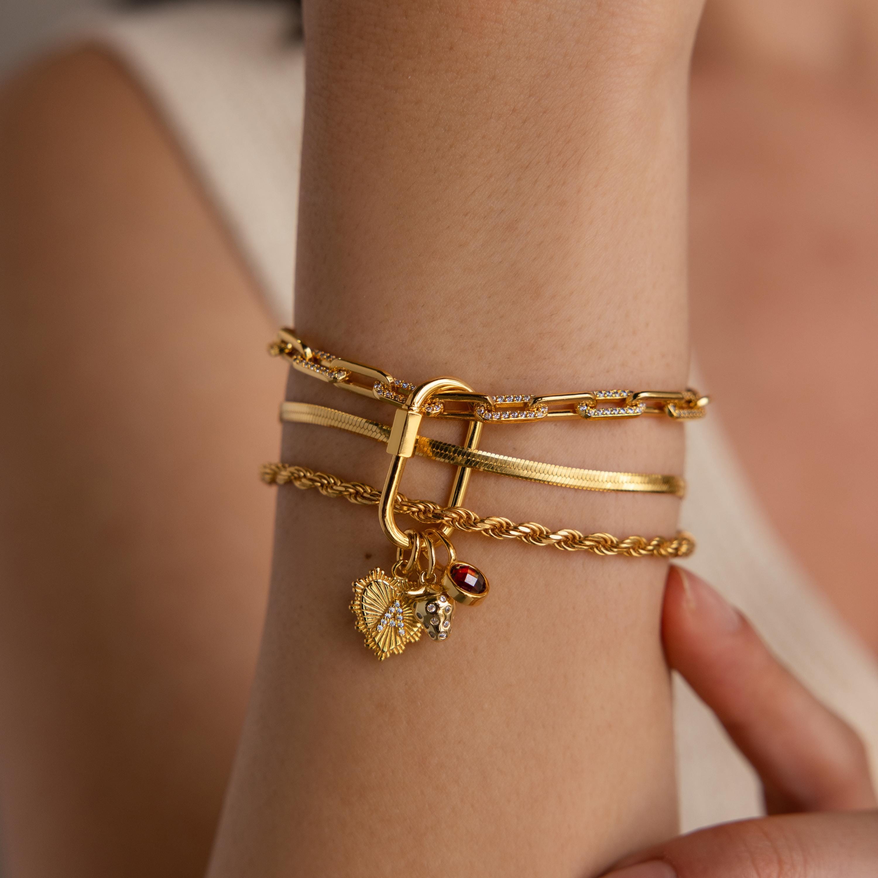 A layered gold bracelet stack on a wrist, featuring the Ribbed Heart Initial Pendant alongside a red gemstone charm and a strawberry charm attached to a carabiner clasp