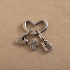 Silver heart-shaped connector styled with three charms: a silver puffed heart, a evil eye hamsa hand, and a silver bubble letter “B,” all arranged neatly on a neutral background.
