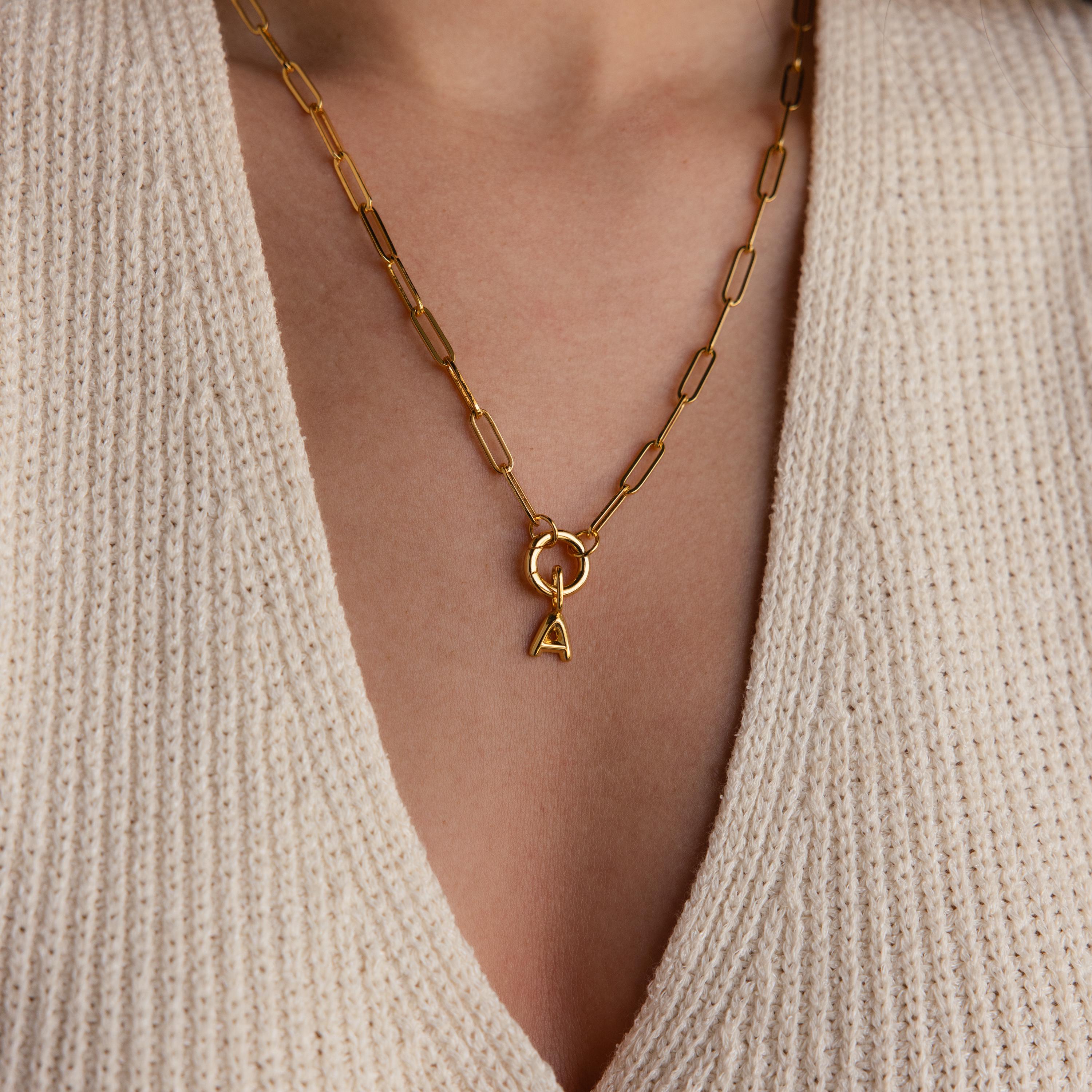 Model wearing a gold paperclip chain necklace with a single bubble letter “A” charm hanging from a Rolo Circle connector, styled over a beige knit top.