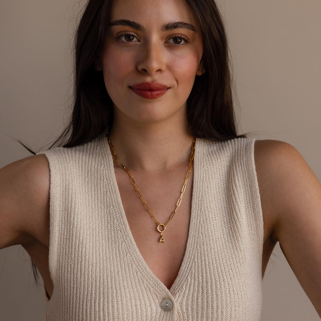 Woman with long dark hair in a cream knit vest wears a gold chain necklace featuring the Bubble Letter Charm, smiling softly at the camera.