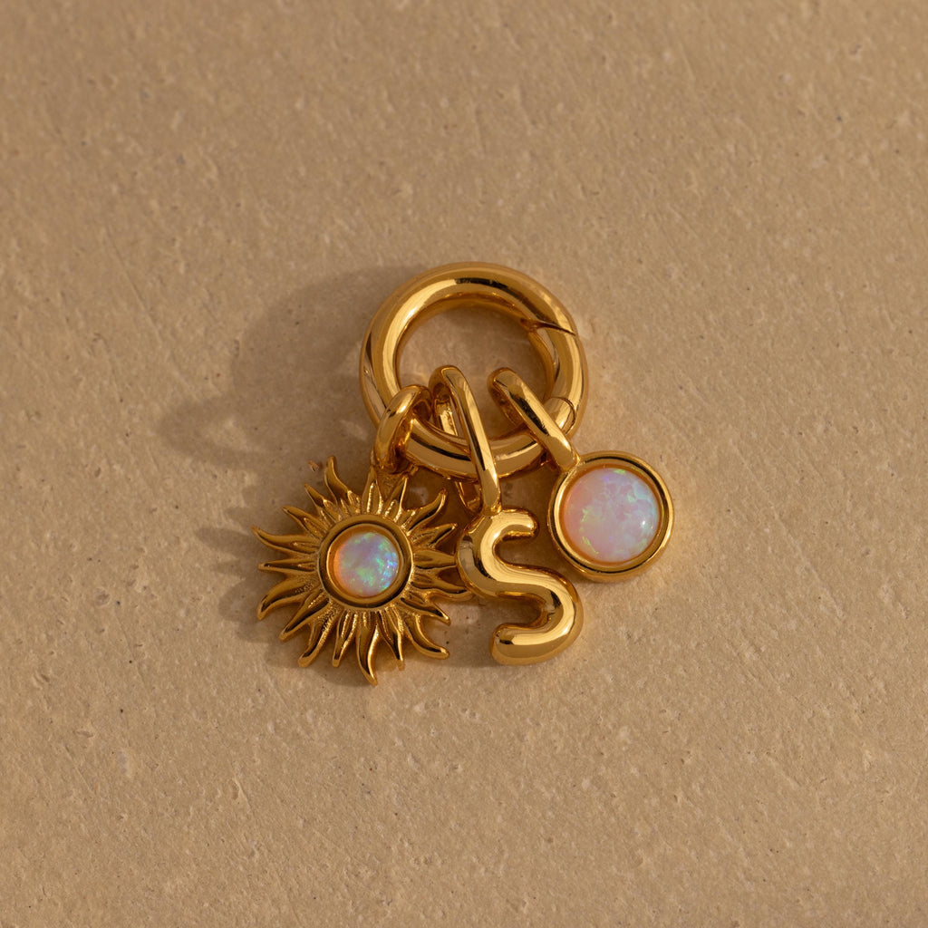 Close-up of three gold charms—an Opal Sun Charm, a bubble letter “S” charm, and a round opal dot charm—hanging together on a polished Rolo Circle connector, displayed on a beige textured surface.