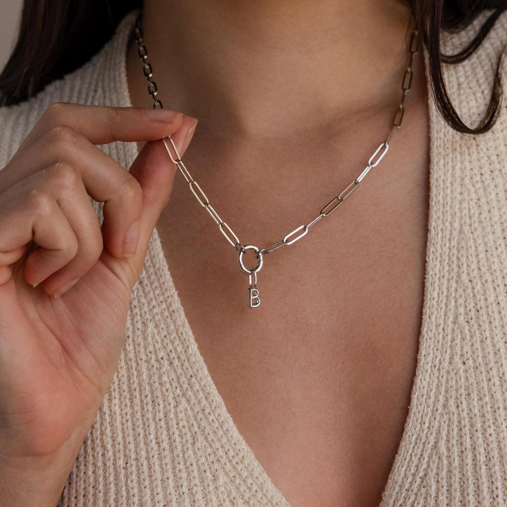 Model holding a silver chain necklace with a bubble letter “B” charm hanging from a silver Rolo Circle connector, styled over a neutral knit top.