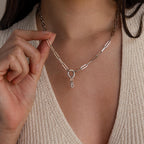 Model holding a silver chain necklace with a bubble letter “B” charm hanging from a silver Rolo Circle connector, styled over a neutral knit top.