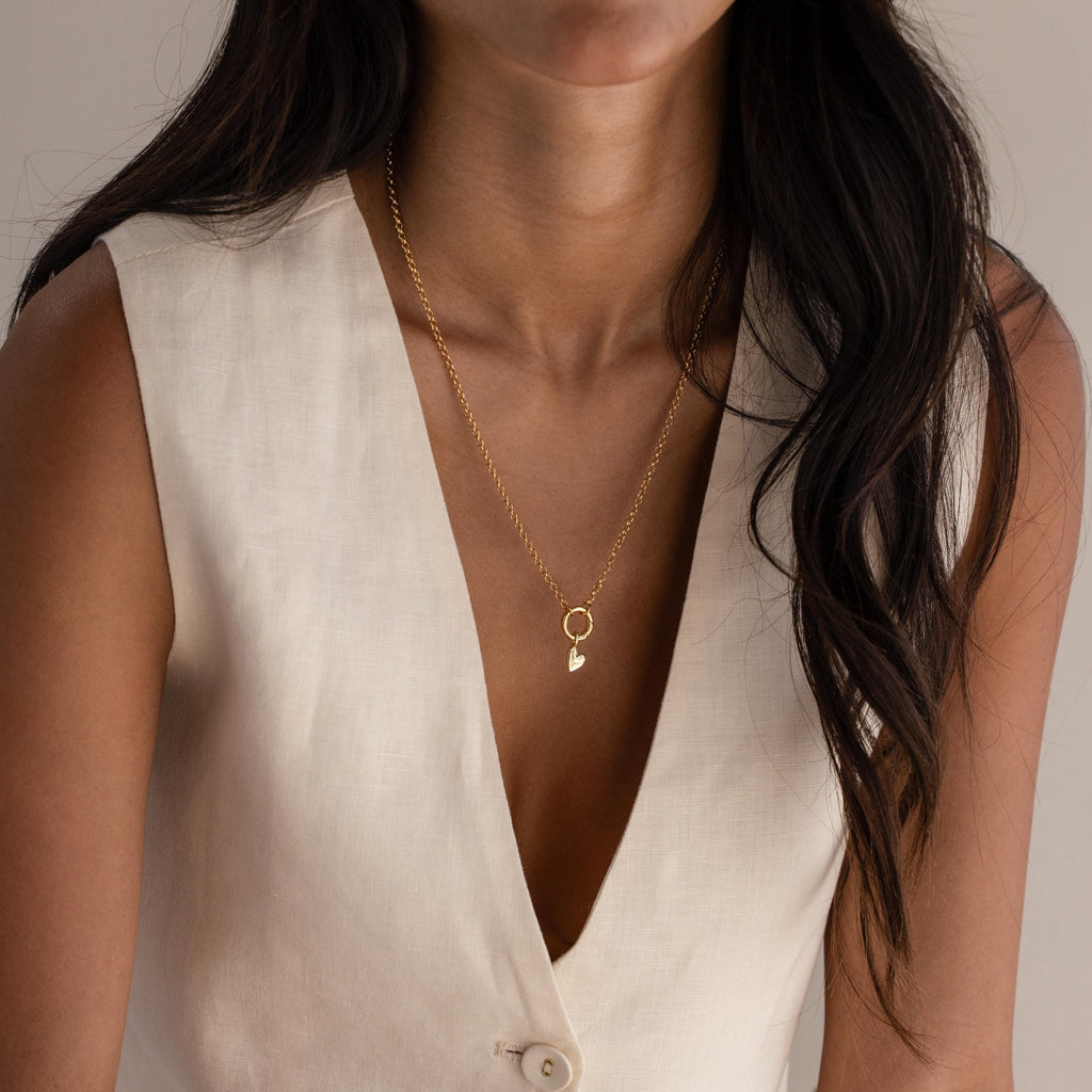 A woman models a minimalist gold chain necklace with a single Simple Heart Charm dangling at the center, styled simply over a deep V-neck blouse.