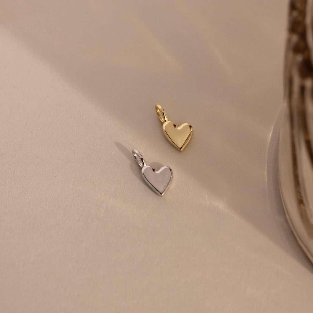 Two Simple Heart Charms, one in silver and one in gold, are placed side by side on a neutral fabric, emphasizing their color and form variations.