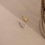 Two Simple Heart Charms, one in silver and one in gold, are placed side by side on a neutral fabric, emphasizing their color and form variations.