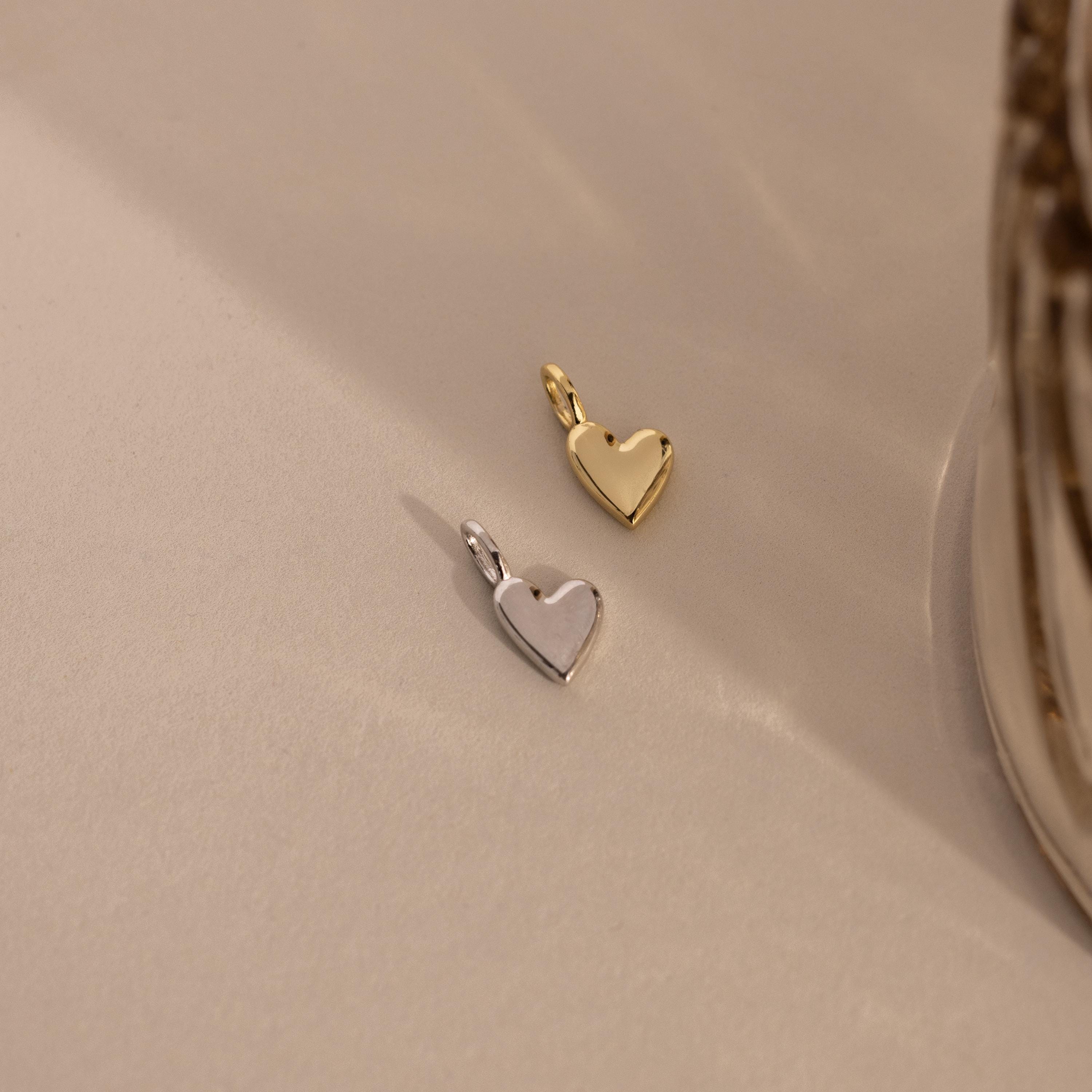 Two Simple Heart Charms, one in silver and one in gold, are placed side by side on a neutral fabric, emphasizing their color and form variations.