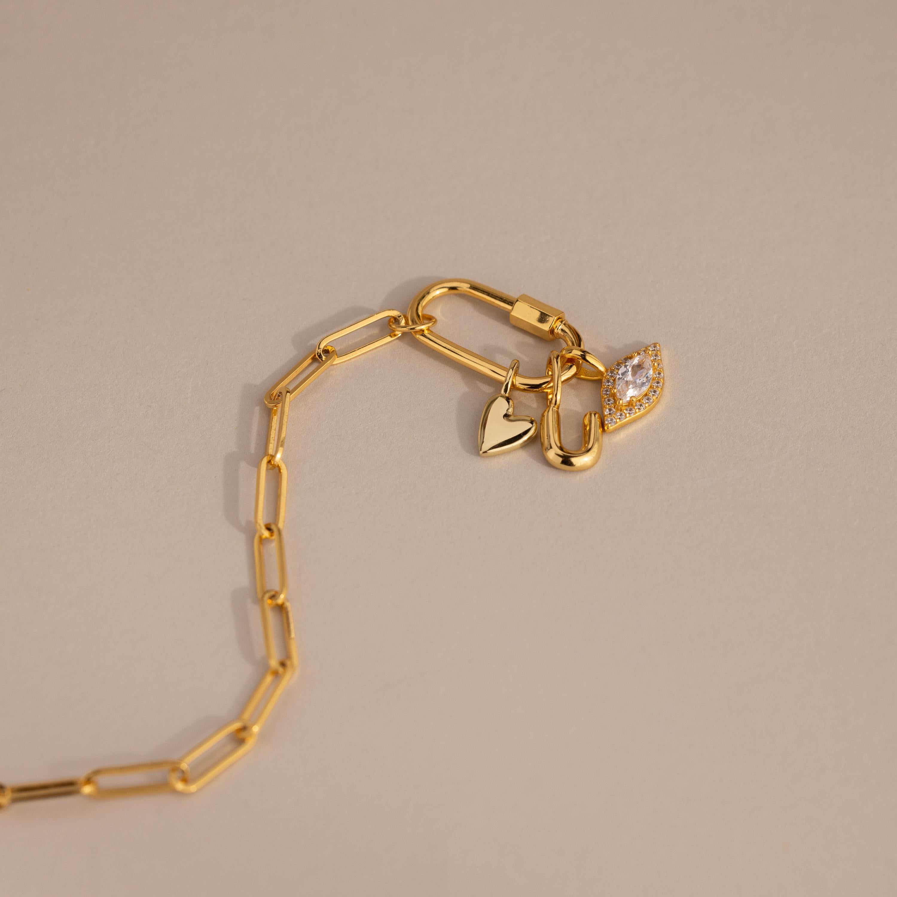 A flat-lay view of a gold paperclip chain featuring a carabiner lock and three attached charms: the Simple Heart Charm, a U-lock charm, and a pavé marquise charm.