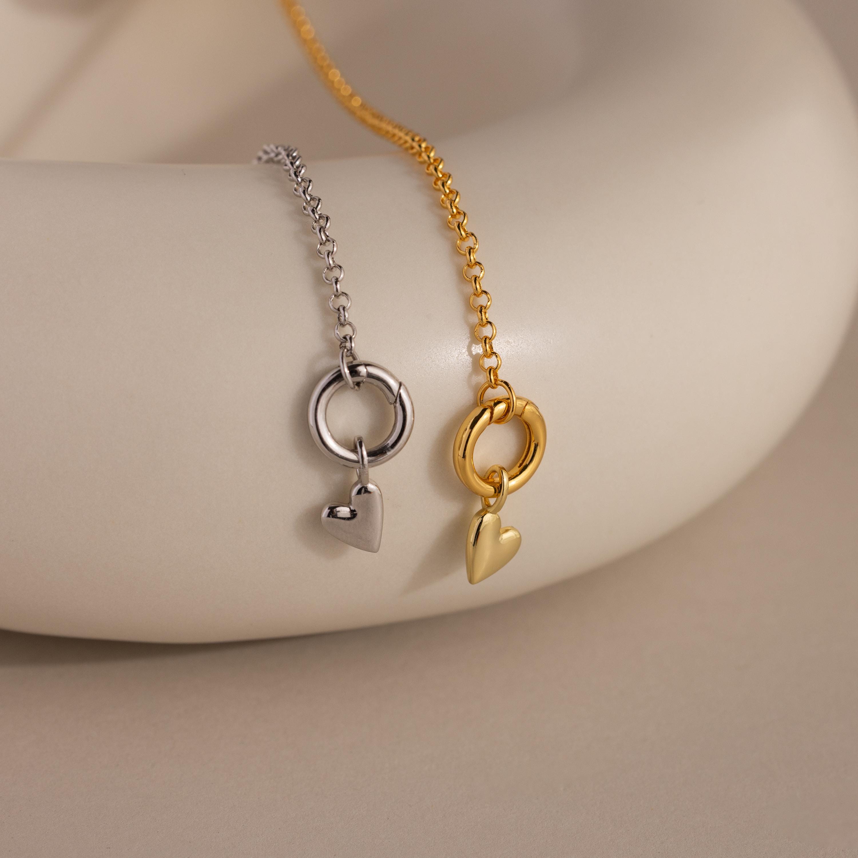 Two Simple Heart Charms in gold and silver finishes are displayed on chain necklaces draped over a smooth, cream-colored ceramic surface.