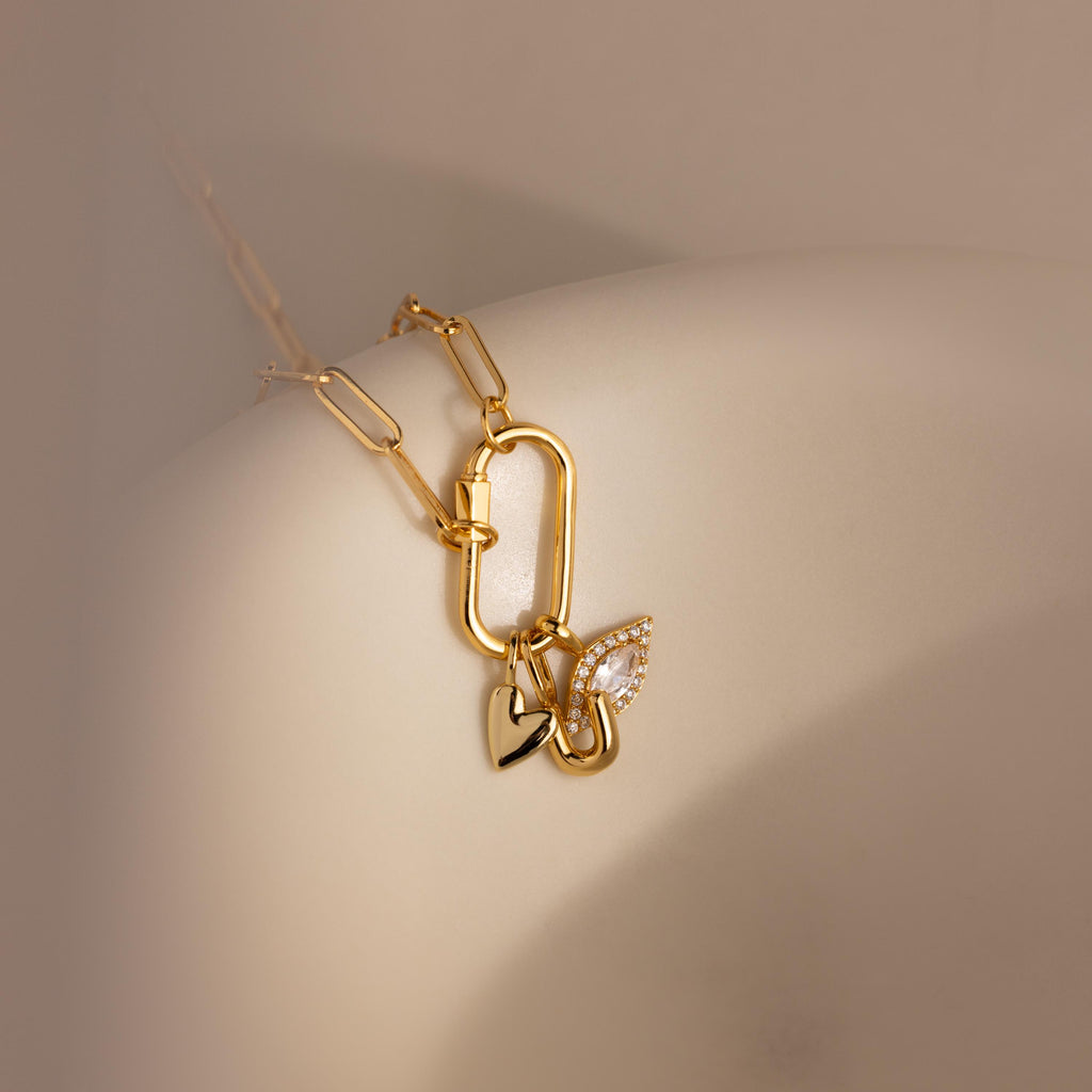 A detailed close-up of a necklace styled with three charms—including the Simple Heart Charm—hanging from a carabiner lock on a curved ceramic form.