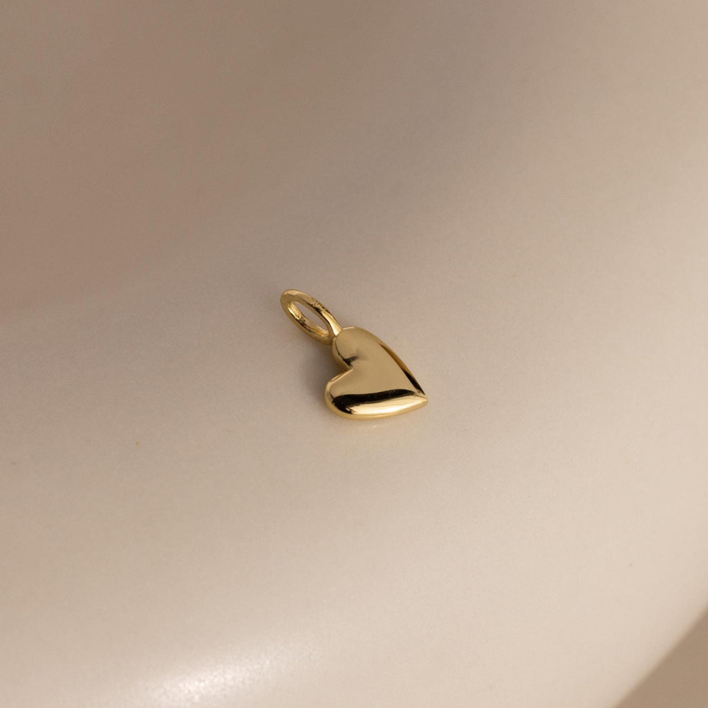 A gold Simple Heart Charm rests on a soft beige backdrop, its curved silhouette and high-polish finish catching the light.