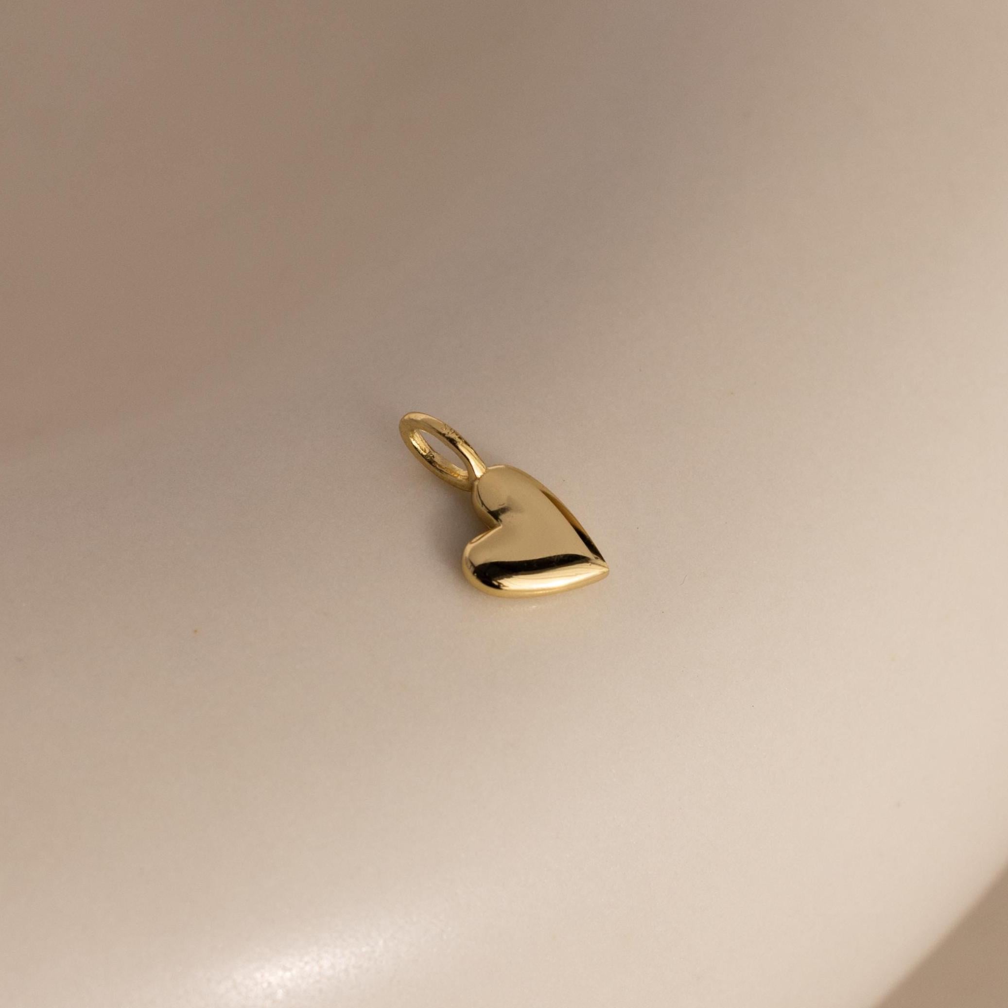 A gold Simple Heart Charm rests on a soft beige backdrop, its curved silhouette and high-polish finish catching the light.