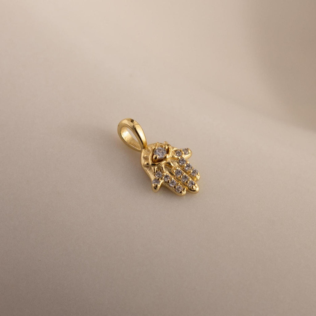 A close-up of the gold Evil Eye Hand Charm lying on a soft beige surface, showcasing its detailed stone-studded design.