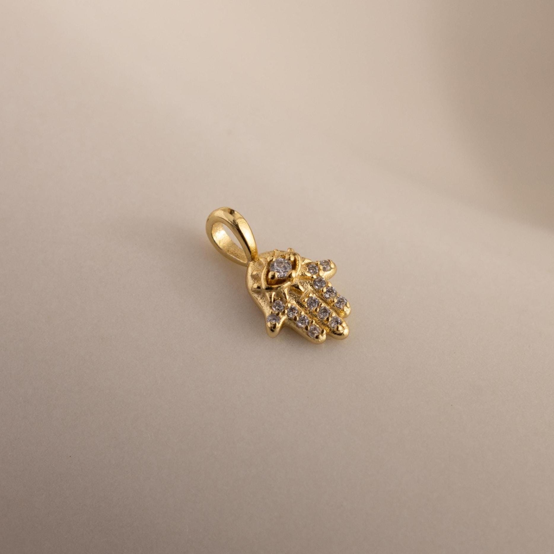 A close-up of the gold Evil Eye Hand Charm lying on a soft beige surface, showcasing its detailed stone-studded design.