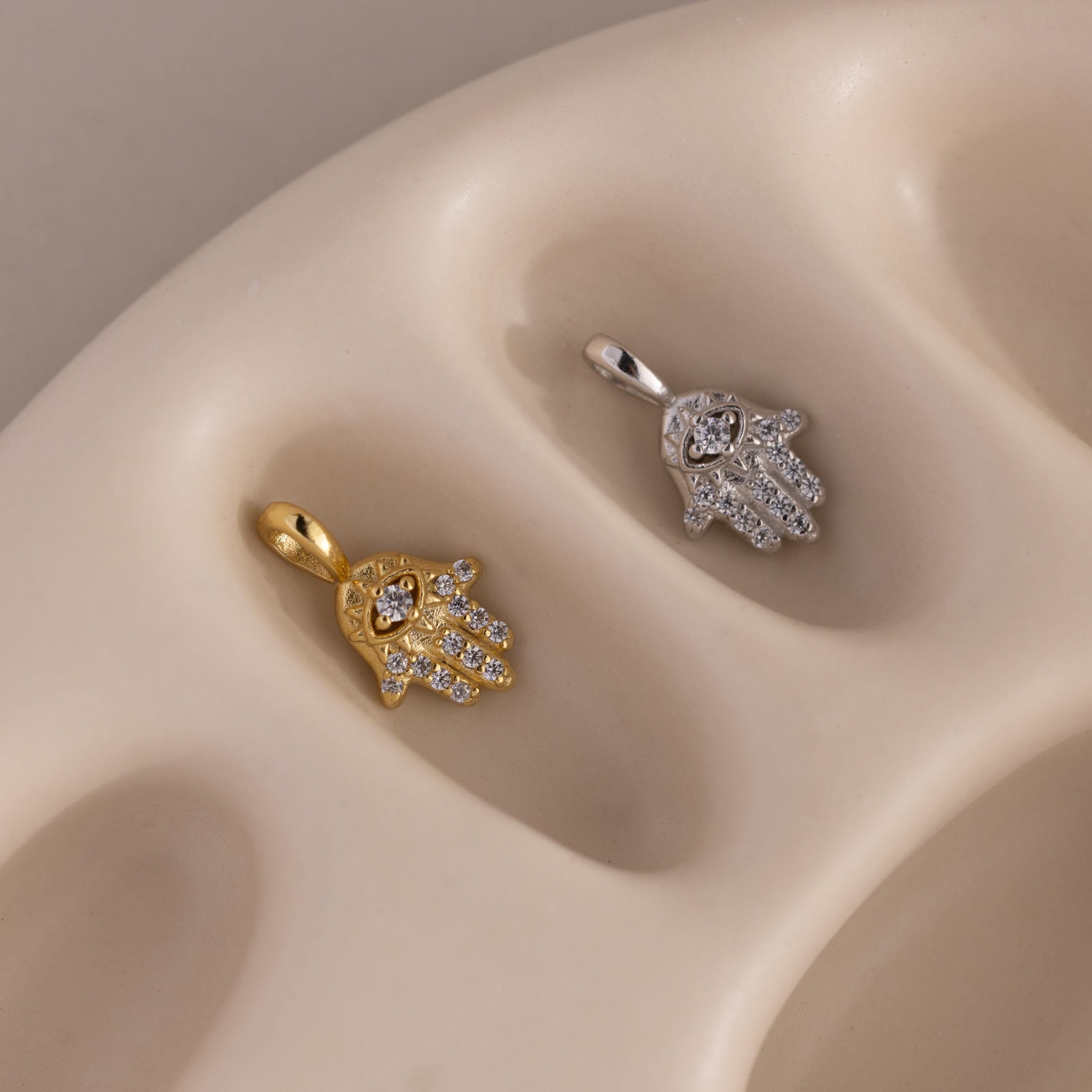A gold and a silver Evil Eye Hand Charm side by side resting in the carved surface of a ceramic jewelry holder.