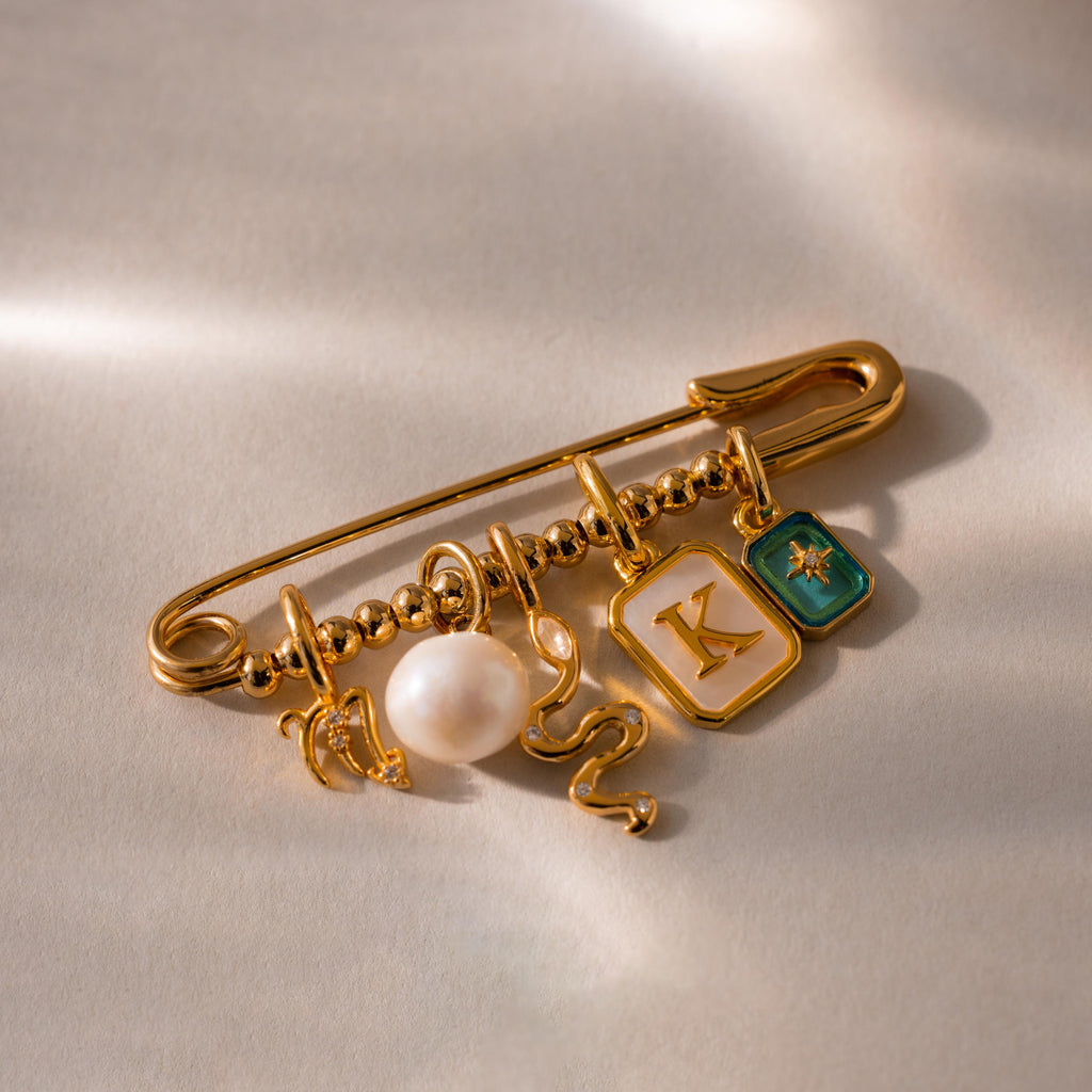 A beaded safety pin charm adorned with various gold charms including a pearl, a snake, and initial K charm on a soft, neutral background.