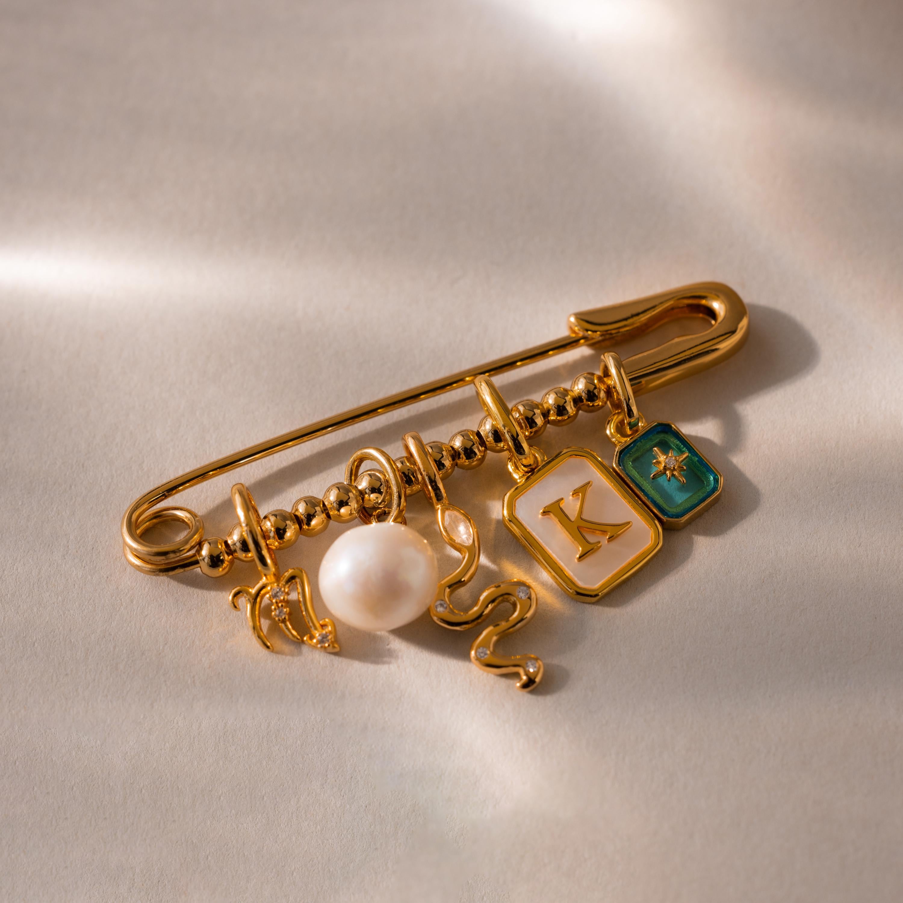 A beaded safety pin charm adorned with various gold charms including a pearl, a snake, and initial K charm on a soft, neutral background.