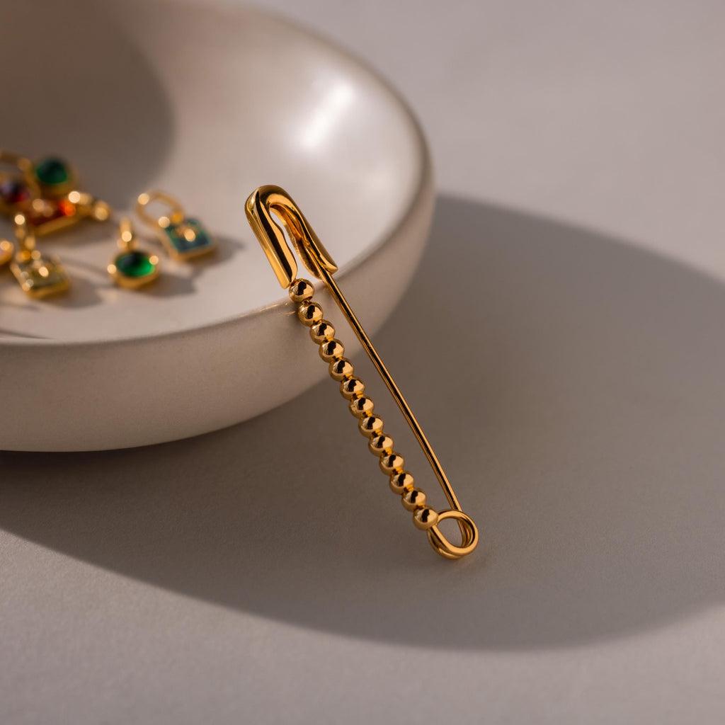 Gold beaded safety pin positioned vertically on a dish with green gemstone charms visible in the background.