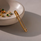 Gold beaded safety pin positioned vertically on a dish with green gemstone charms visible in the background.