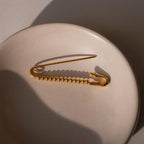 A gold safety pin with round beaded detailing laid flat on a ceramic dish, displayed without charms.
