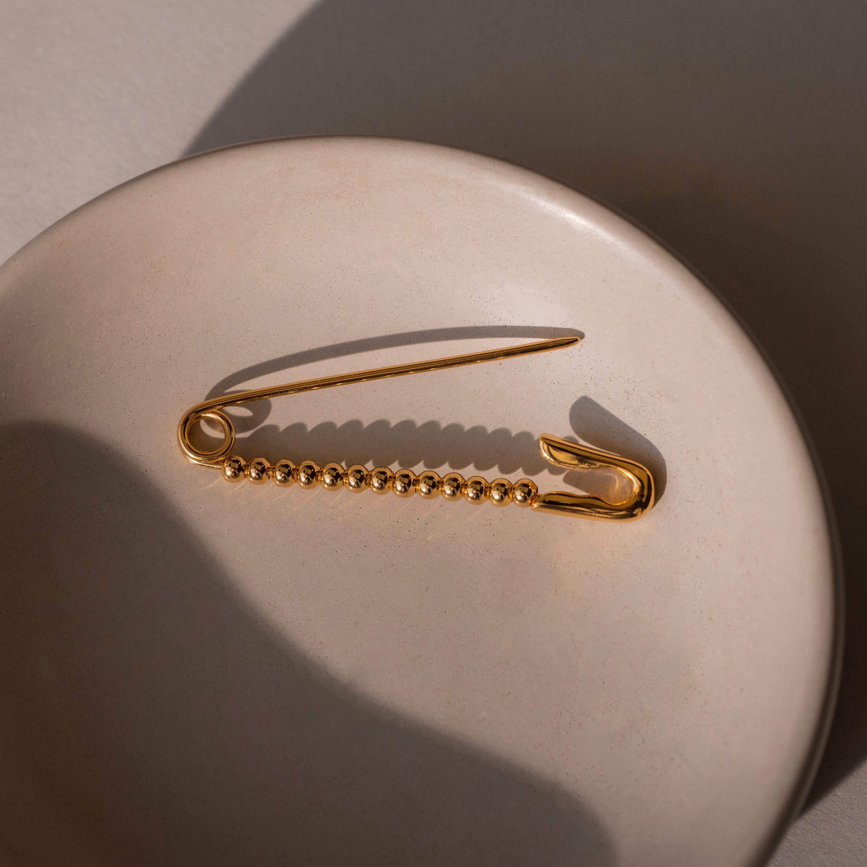 A gold safety pin with round beaded detailing laid flat on a ceramic dish, displayed without charms.