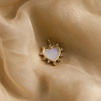 The gold Pearl Starburst Heart Charm rests on a soft, golden fabric surface, highlighting its iridescent center and sparkle.