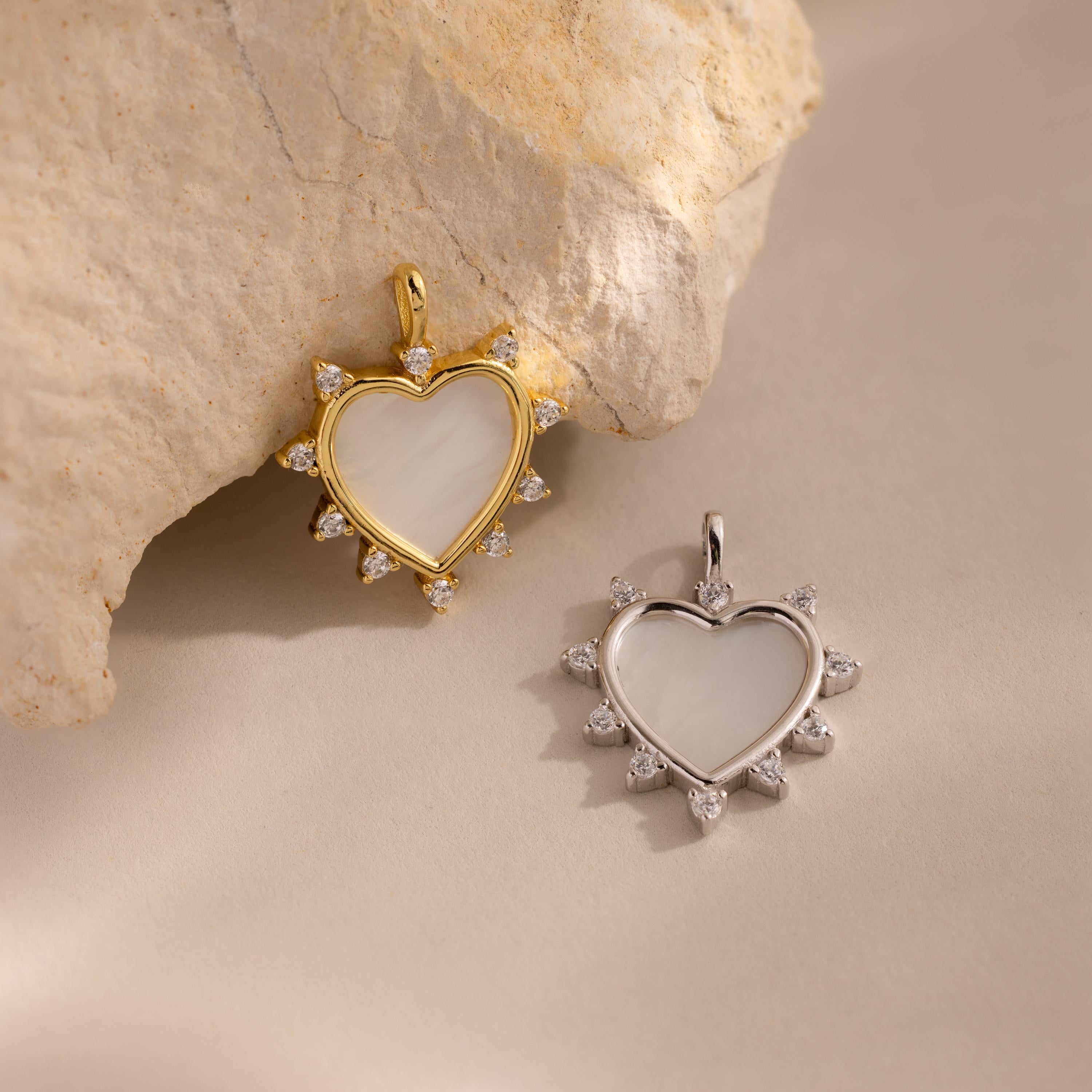 A close-up image of two heart-shaped Starburst Pearl Heart Charms displayed on a matte surface. The charm on the left features a gold-plated setting, with a luminous mother-of-pearl heart center bordered by ten sparkling cubic zirconia stones arranged in a radiant starburst pattern. The charm on the right is the same design in silver