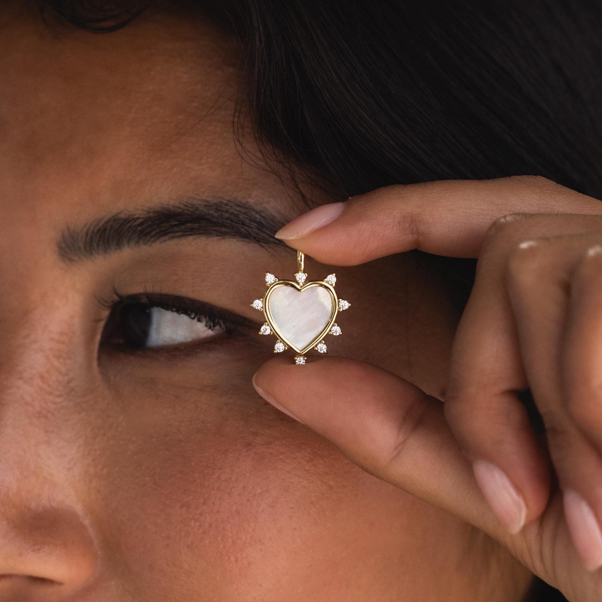 A person holds the gold Starburst Pearl Heart Charm delicately between two fingers in front of their face, showcasing the charm's size and radiance.