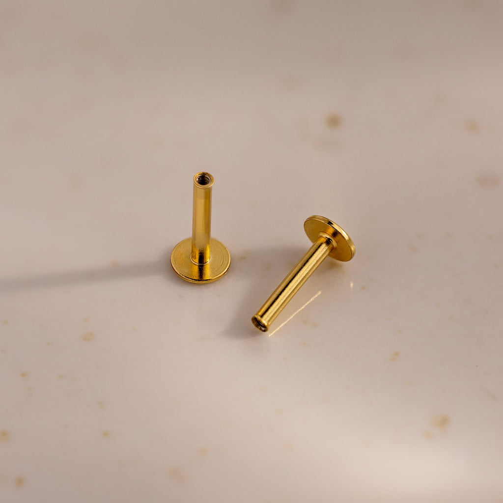 A pair of 18k gold Flat Back Posts positioned on a speckled ceramic surface, one standing and one lying down.