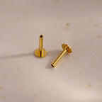 A pair of 18k gold Flat Back Posts positioned on a speckled ceramic surface, one standing and one lying down.