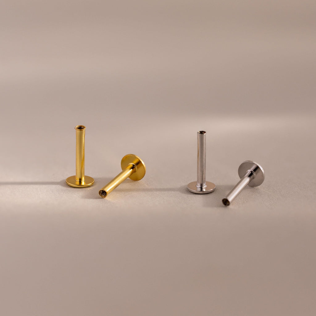 A set of four flat back posts, two in a warm 18k gold finish and two in a cool sterling silver finish, are elegantly arranged on a soft matte surface. Each post features a smooth cylindrical stem with a threaded interior, designed to securely hold jewelry tops. The bases are circular and flat, providing stability and comfort