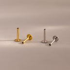 A set of four flat back posts, two in a warm 18k gold finish and two in a cool sterling silver finish, are elegantly arranged on a soft matte surface. Each post features a smooth cylindrical stem with a threaded interior, designed to securely hold jewelry tops. The bases are circular and flat, providing stability and comfort