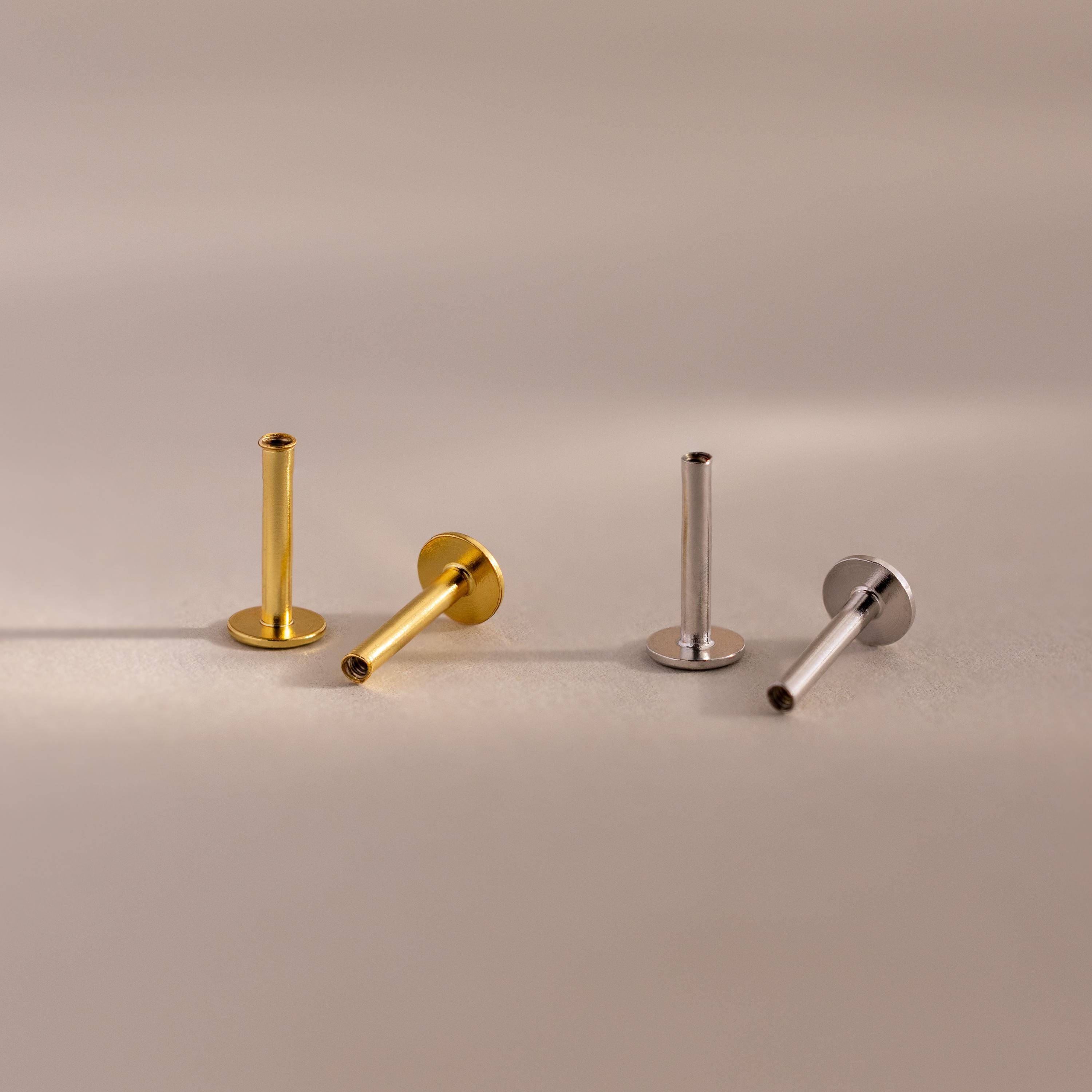 A set of four flat back posts, two in a warm 18k gold finish and two in a cool sterling silver finish, are elegantly arranged on a soft matte surface. Each post features a smooth cylindrical stem with a threaded interior, designed to securely hold jewelry tops. The bases are circular and flat, providing stability and comfort