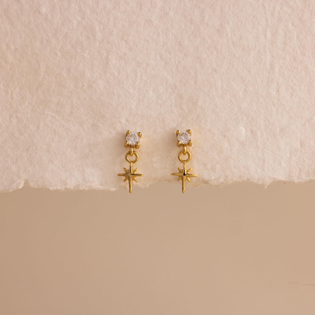 A straight-on product display of the 18k gold Diamond Star Flatback Earrings against a soft beige and off-white background, highlighting the charm's clean symmetry.