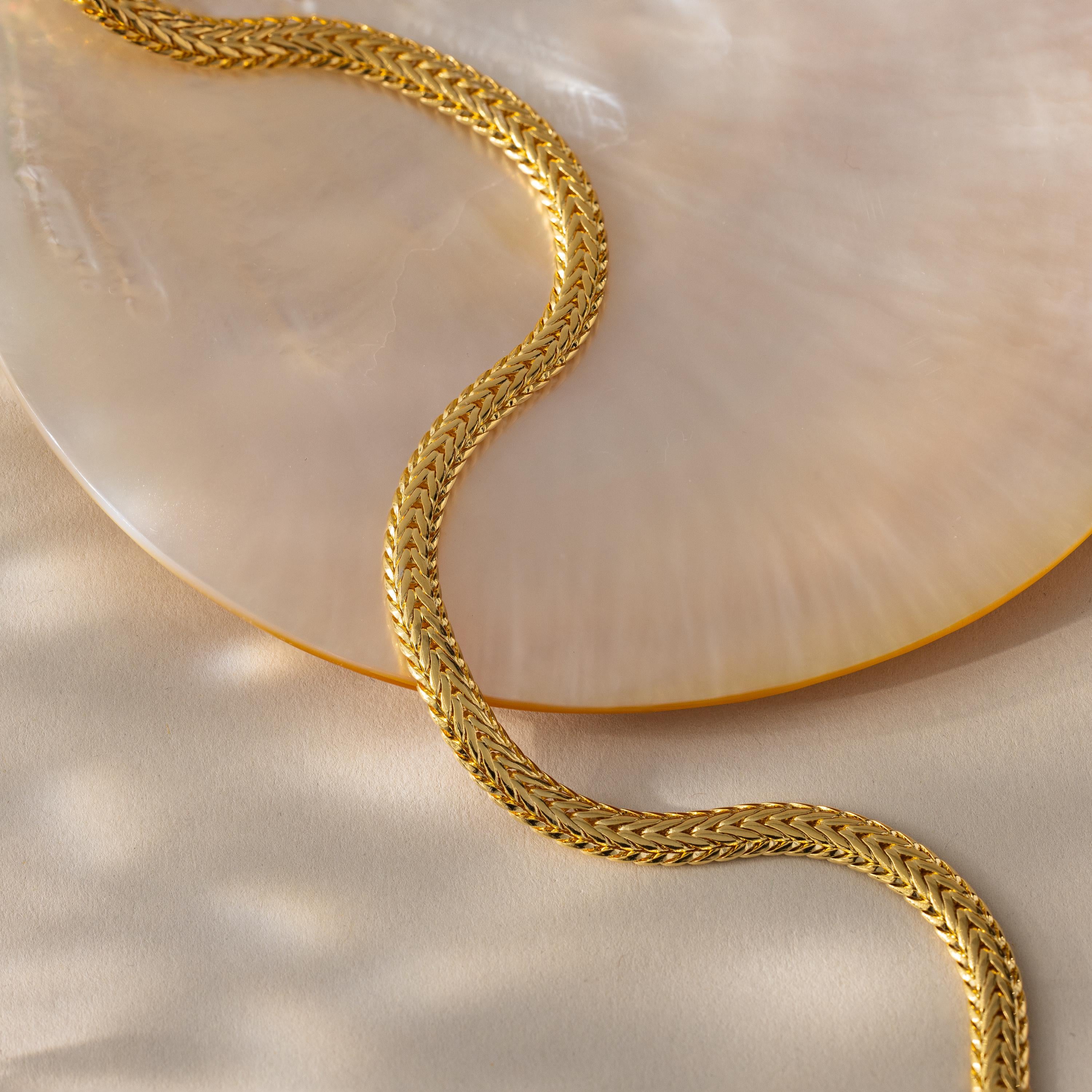 A single gold Foxtail Chain Necklace curves gracefully on a flat cream-colored surface beside a polished shell dish