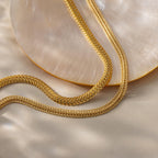 A  gold Foxtail Chain Necklace and a Mesh Chain Necklace styled in a flowing “S” curve across a neutral beige surface next to a polished, iridescent shell
