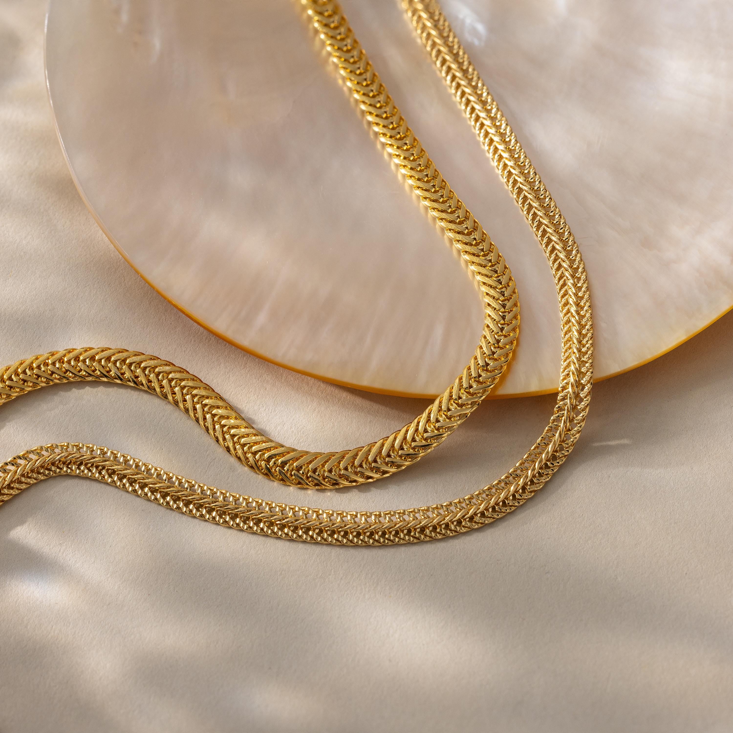 A  gold Foxtail Chain Necklace and a Mesh Chain Necklace styled in a flowing “S” curve across a neutral beige surface next to a polished, iridescent shell
