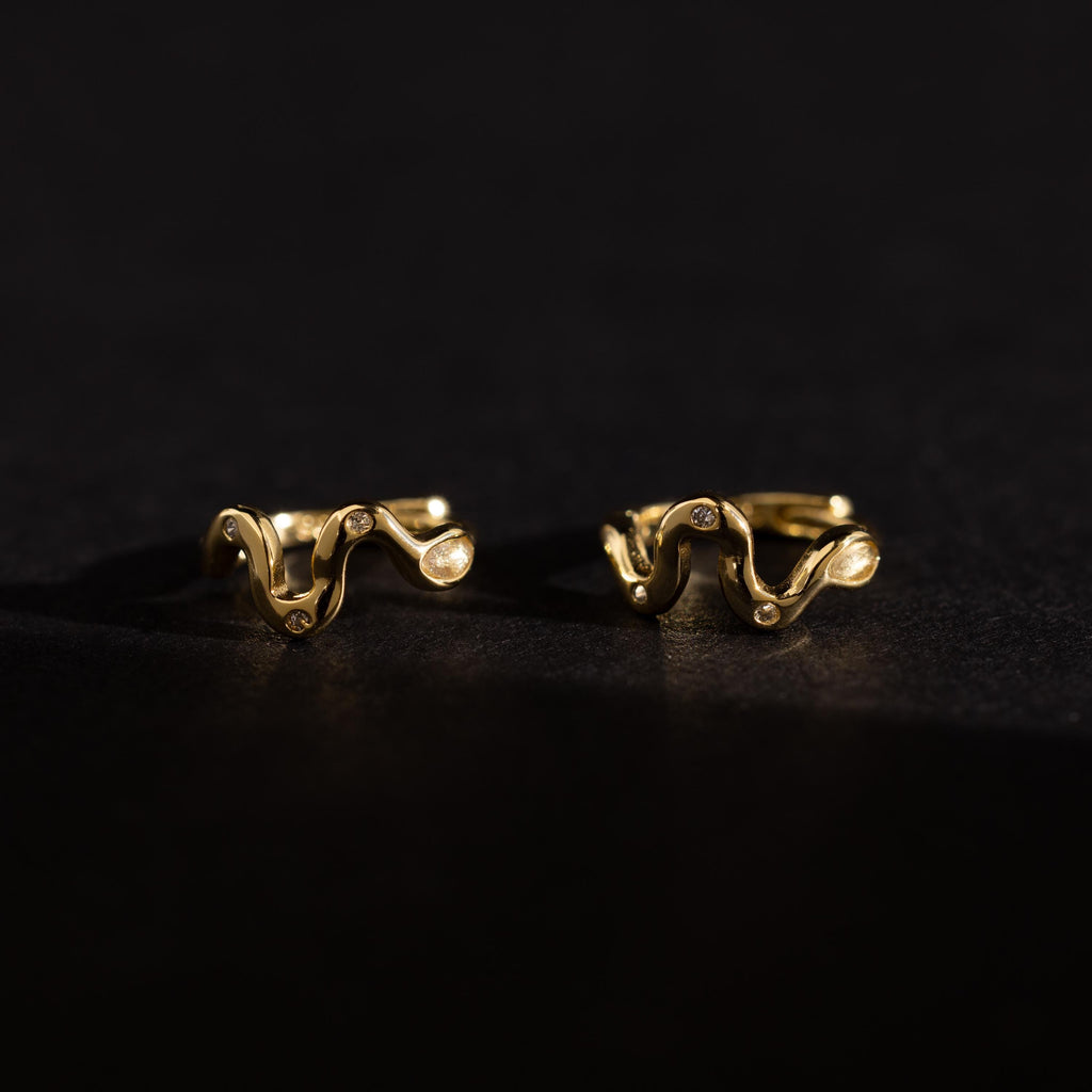 Pair of Pavé Viper Huggie Earrings in gold displayed on a dark surface, showcasing their wavy shape and pavé accents.