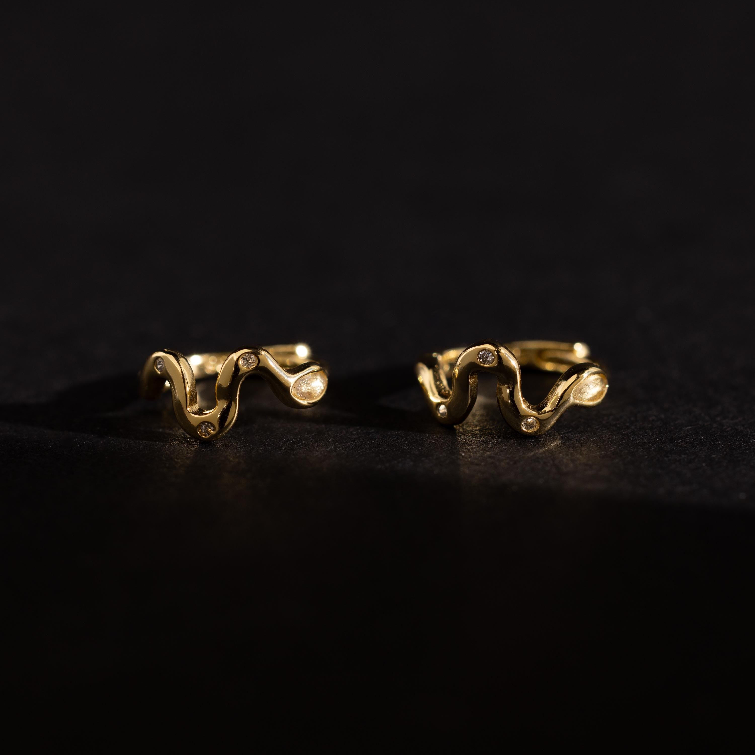 Pair of Pavé Viper Huggie Earrings in gold displayed on a dark surface, showcasing their wavy shape and pavé accents.