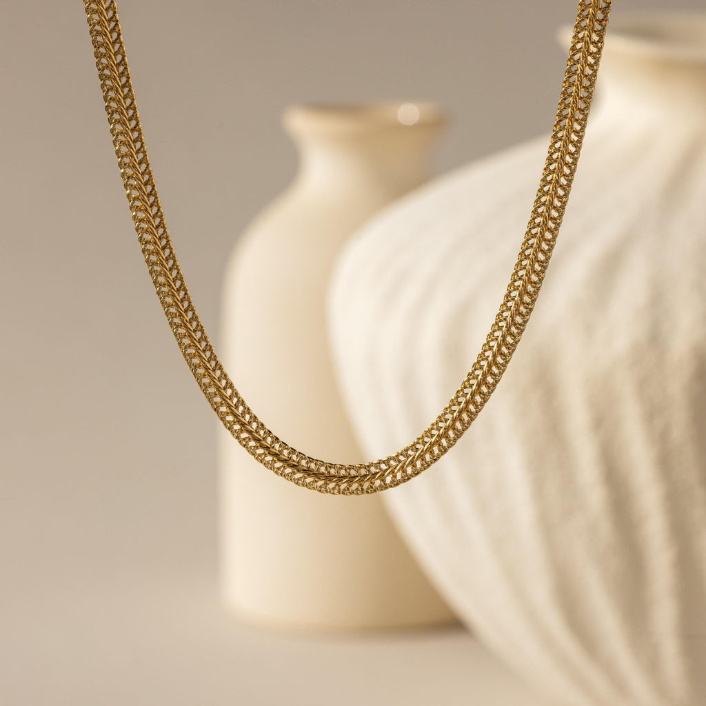 hick Mesh Chain Necklace in gold floats against a soft neutral backdrop. The lighting enhances the tightly interlocked mesh links, showcasing the necklace’s uniform weave