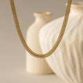hick Mesh Chain Necklace in gold floats against a soft neutral backdrop. The lighting enhances the tightly interlocked mesh links, showcasing the necklace’s uniform weave