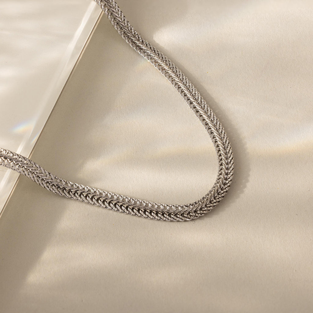 Close-up of a silver mesh chain necklace with intricate woven texture, displayed on a neutral background with soft lighting.