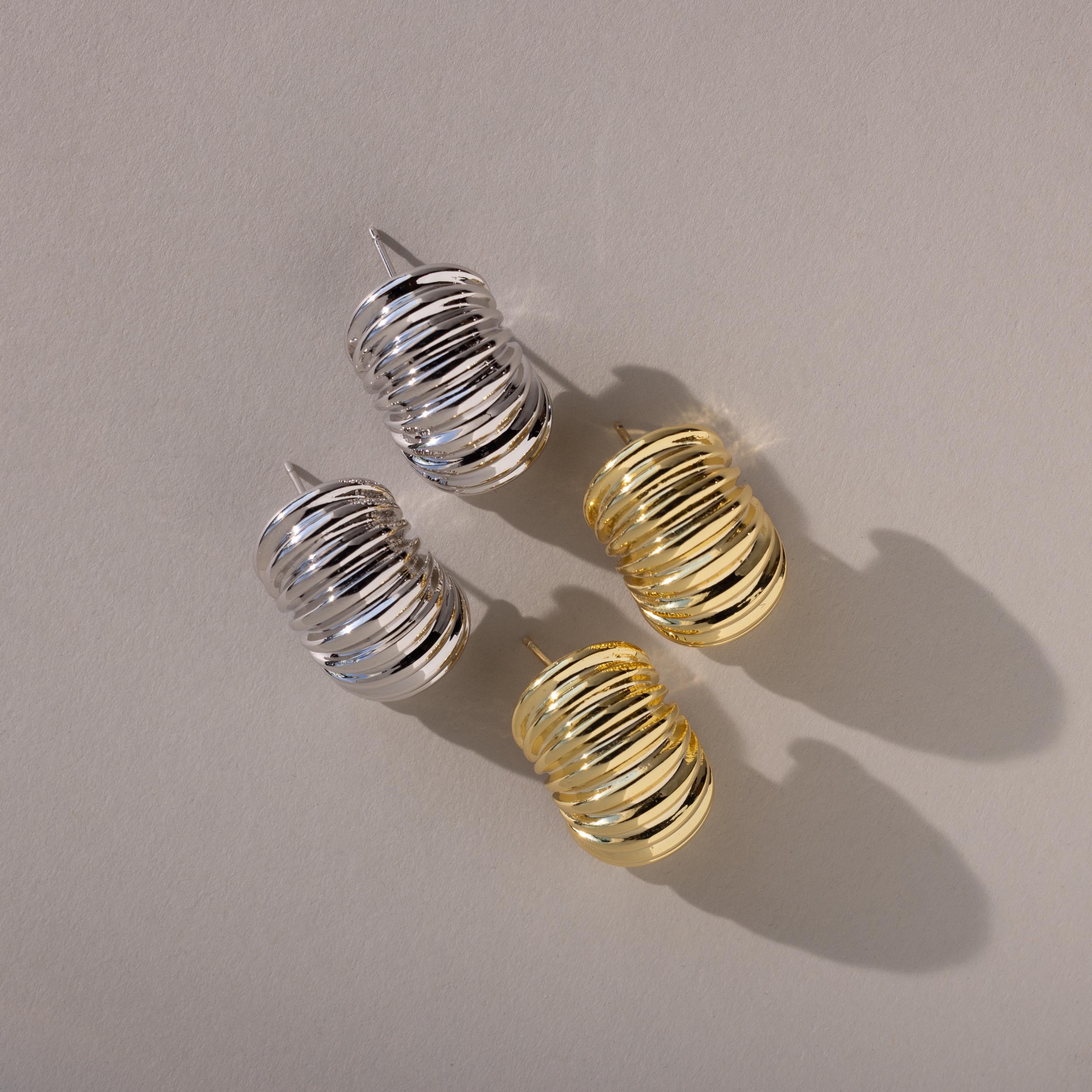 Aerial view of two pairs of chunky ribbed earrings—one in silver, one in gold—placed neatly with their posts visible and shadows cast beneath.