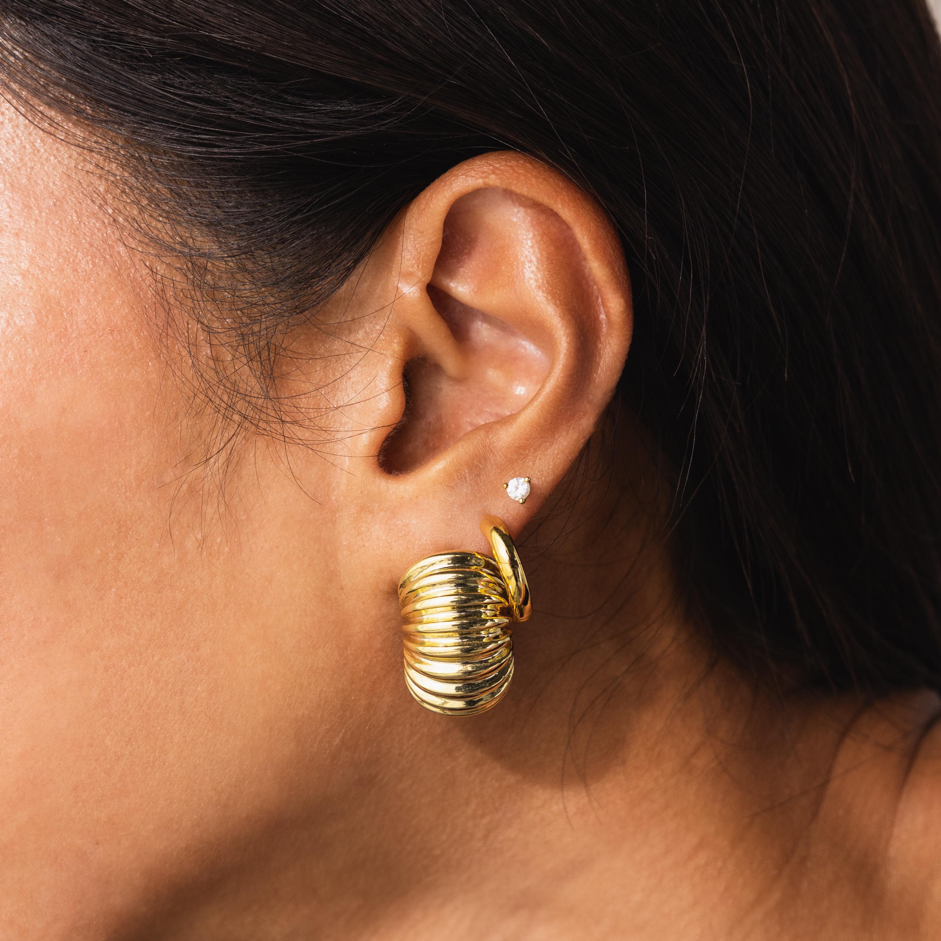 Model wearing the chunky gold ribbed earring paired with a small diamond stud and oval hoop above it; the layered look adds dimension to the ear styling.