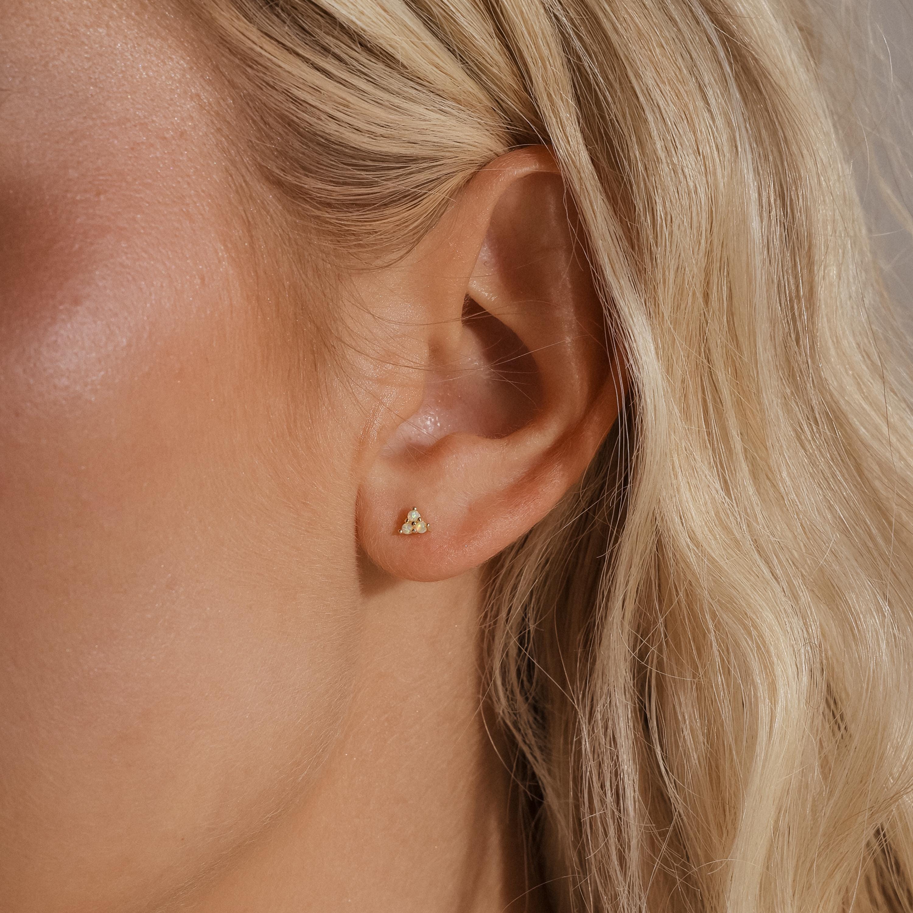 A close-up of a woman's ear showing a single gold Opal Cluster Stud Earring worn on the lobe, capturing its small, triangular shape and opal shimmer.