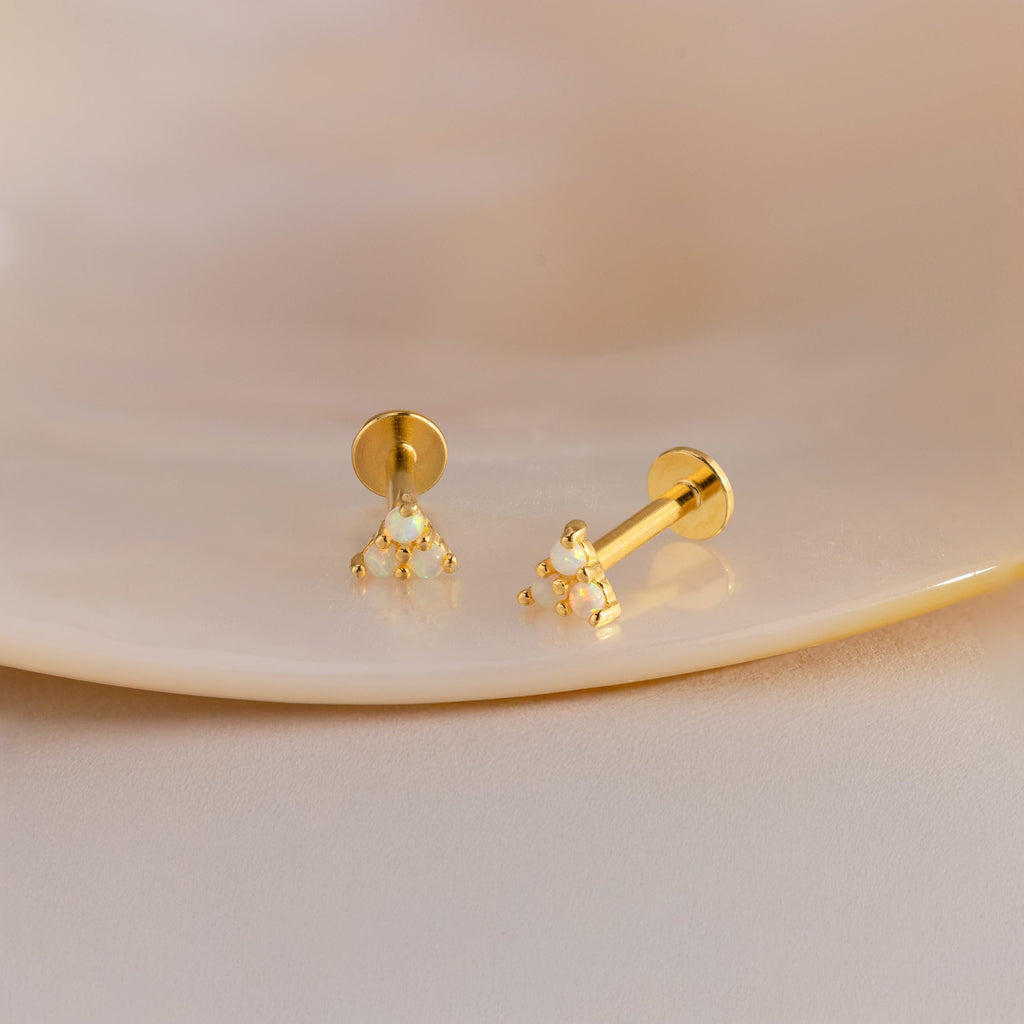 A close-up photograph displays a pair of dainty gold stud earrings resting on a smooth, neutral-toned ceramic surface. Each earring features a triangular cluster of three tiny round opals, each held in place with polished gold prongs. The iridescent opals reflect hues of green, blue, and pink, subtly catching the light.